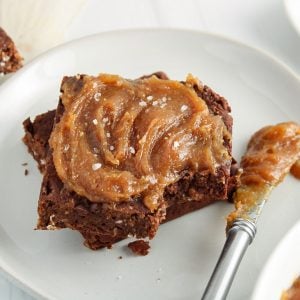 Close up on a piece of vegan brownies that's covered with some homemade peanut butter frosting. There is a silver knife covered with more frosting on the side and more brownies in the background.