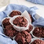 Front view of 9 beetroot chocolate muffins that are in a oval container that's covered with a blue hand towel. You can see empty glasses and a jar filled with vegan milk in the background.