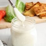 There is a jar that contains a quick cashew mayo with a butter knife taking some to show the texture. In the background, there are the fixings to make a sandwich: roasted tofu, sliced tomatoes, sliced bread and lettuce.