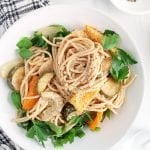 Showing is a white bowl that contains pasta with hummus and roasted vegetables. There are fresh herbs topping the pasta dish. On the side of the bowl, there are sliced lemon, salt and pepper in a small bowl, a glass of water, fresh basil and a black and white hand towel.