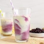There is a tall glass filled with a blueberry pineapple smoothie in a galaxy pattern. There are more fruits in the background as well as a second glass of smoothie. The 2 glasses of smoothies have a straw in them.