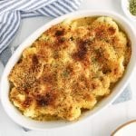 Showing is an oval white dish filled with a vegan cauliflower gratin with its crusty topped browned. There are small bowls on the side with spices, a large serving spoon and a white and blue hand towel.