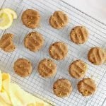 There are a few vegan lemon cookies that are cooling down on a cooling rack. The cookies are sprinkled with lemon zest. There are slices of lemon around the cookies as well as a cup of tea and a yellow hand towel.