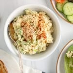 Showing is a small white bowl that contains a vegan eggless salad topped with paprika. The bowl is surrounded with tomatoes, lettuce, toasted bread and open sandwiches made with the salad.