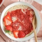 There is a white bowl that contains a creamy barley breakfast bowl that's topped with a strawberry and rhubarb quick compote. There is also a wooden spoon on the side of the bowl and fresh strawberries. Also beside the bowl, there is another breakfast bowl as well as a few small bowl containing more of the compote and fresh strawberries. The bowls are on a large pink hand towel and there is a cup of tea on the side.
