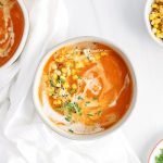There are 2 white bowls that contain a butternut squash and red pepper soup that's topped with coconut milk, corn, chives and black pepper. There are a few bowls around containing more of the toppings.