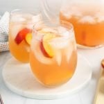 Showing is a glass of Oolong peach iced tea that's filled with ice cubes and slices of peach. There is a large pitcher in the background with more of the tea as well as a second glass with some of the tea. There are pieces of peach on the side and a black and white with dots hand towel on the side.