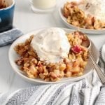 Close up on a vegan rhubarb crumble that's topped with vegan ice cream and is on a small grey plate. There is a spoon on the side of the plate and you can see another plate with more of the crumble that's in the background. Also showing is a blue and grey hand towel that's beside the plates and the large rectangular baking dish containing the crisp.
