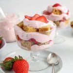 Front view on a mini vegan strawberry trifle that layers cubes of cake, a pink aquafaba strawberry mousse and fresh sliced strawberries. There are more mini trifle in the background as well as spoon and fresh strawberries.
