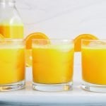 Showing are 3 glasses containing ginger turmeric shots with a slice of orange on the side of the glass. There is another large juice container in the background.