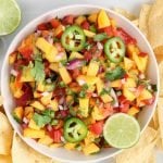 Overhead view on a white bowl that's on a large white serving plate. The bowl contains a homemade spicy jalapeño salsa and the plate has tortilla chips surrounding the salsa bowl. There are a few fresh limes also around on the plate.