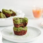 Front view on 2 mini matcha brownies that are pilled on top of each other. You can nicely see their bottom dark layer and top bright green layer. These 2 brownies are on a white plate and the top mini brownie has a bite taken off. You can see more of them in a bowl in the background. Also in the background, there are 2 cups of tea and a matcha sifter with some powder inside that's on a plate.