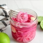 Front angle view on a jar containing Mexican pickled onions. The on ion are bright pink and you can see a fork holding some of the onion to show the texture. There are also halved limes in the background.