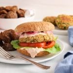 Showing is a vegan chickpea and oat burger that's on a plate with a side of roasted potatoes. The burger also layers tomatoes, lettuce, a pinkish sauce and pickled red onions. You can see more roasted potatoes and burger patties in the background, a jar with some more pickled onions as well as a blue hand towel.