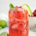 Close up on a virgin strawberry mojito. You can see the texture through out the glass made with crushed strawberries and mint. There are more fresh lime, strawberries and mint in the background.