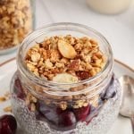 Front angle view on a glass jar that layers a chia pudding, with chopped fresh cherries and topped with an homemade vanilla almond granola. You can see more granola in a big jar in the background as well as more cherries.