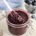 Front angle view on a jar filled with an easy blueberry chia jam. There is a small white spoon taking some of the jam to show the texture and you can see some fresh blueberries, chia seeds and a blue hand towel in the background.
