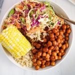 Overhead view on a bowl that contains brown rice, corn on the cob, a vegan creamy coleslaw and some BBQ chickpeas. There is a fork on the side of the plate.