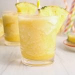 Front view on a short glass containing a homemade pineapple slush. There is a yellow straw in the glass and also a wedge of pineapple against the rime of the glass. You can see more glasses of the slush in the background as well as more straw and fresh fruits.