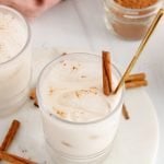 Close up on a short glass that's filled with ice cubes and a homemade brown rice vegan horchata. There is a tall gold spoon in the glass as well as a cinnamon stick. There are more cinnamon sticks on the table as well as a small bowl with ground cinnamon to sprinkle over the glasses.