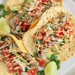 Close up on 3 vegan fish tacos that on a white plate. You can see their filling consisting of vegan fish made with hearts of palm, pico de gallo, avocado slices and a green cabbage made with lime and cilantro.