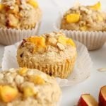 Front angle view (close up) on a vegan peach muffin that on a large white plate. The muffin still is on its muffin tin and there are more muffins around as well as a few slices of peach.