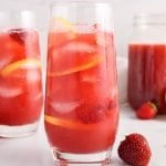 Front view on a tall glass filled with a strawberry acai lemonade refresher. You can see fresh strawberries, ice cubes and lemon slices within the lemonade. There is also a second glass of lemonade in the background as well as a larger pitcher with more of it.