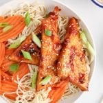 Overhead view on a sticky tofu that's topping a bowl filled with rice noodle and steamed carrots. The tofu is also garnished with white sesame seeds and chopped green onion. There are small bowls on the table containing more sesame seeds and chopped green onions.
