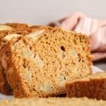 Front view on a half sliced vegan apple cinnamon bread. There are fresh apples, sliced apples and a pink hand towel in the background. The focus is on one of the slices of apple bread to show the texture and you can see all the diced apple within the slice.