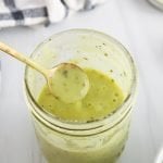 Front angle view on a jar that contains a vegan Italian dressing. The sauce has a light green color and you can see a black and white hand towel, a large shallow bowl with a salad and 2 cherry tomato on the table. You can see someone holding a small spoon that's covered with some of the dressing that's dripping back in the jar to show the texture of the salad vinaigrette.