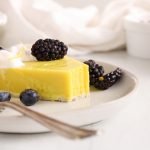 Front angle view on a slice of vegan lemon pie that's placed on a plate and garnished with berries and vegan whipped cream. There is a fork with some pie on it also on the plate.