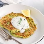 Front angle view on 2 vegan zucchini basil fritters placed on a small plate and garnished with a vegan dill yogurt sauce. There is a lemon wedge on the side and the fritters are garnished with ground black peppers.