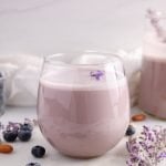 Front view showing a glass on blueberry lavender almond milk. You can see a few lavender flowers floating on top of the milk, more fresh blueberries in the background and a larger jar filled with more of the milk in the background as well.