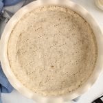 Overhead view on a gluten-free shortcrust pastry that's left empty still in the pie plate. The pie crust is on a blue towel and there are 2 bowls on the side containing ground chia seeds and almond flour.