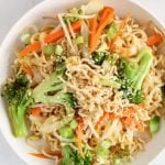 Overhead view on a white bowl containing a vegan chow mein recipe. You can see a close up on the noodles, broccoli, carrots and bean sprouts and there is a garnished of sesame seeds over the bowl.
