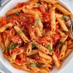 Overhead view on a bowl containing a penne pomodoro pasta dish. The noodles are also garnished with vegan parmesan cheese and more fresh basil. There is a fork in the side of the bowl, a black and white hand towel and a small bowl with more parmesan cheese.