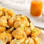 Front angle view on a pile of spicy orange cauliflower bites that are on a large rectangular white plate. There is a large serving spoon on the side as well as more orange sauce and sesame seeds in the background.