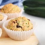 Close up on a vegan zucchini muffin still in his paper tin and placed on a wooden board. There are fresh zucchinis in the background as well.