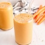 Front angle view on a tall glass that's filled with a homemade banana carrot smoothie. There are some carrots in the background, bananas, a small bowl with hemp seeds.