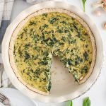 Overhead view on a vegan spinach quiche with a slice that was taken off. There are on the side a hand towel, garlic, nutritional yeast in a bowl and black pepper.