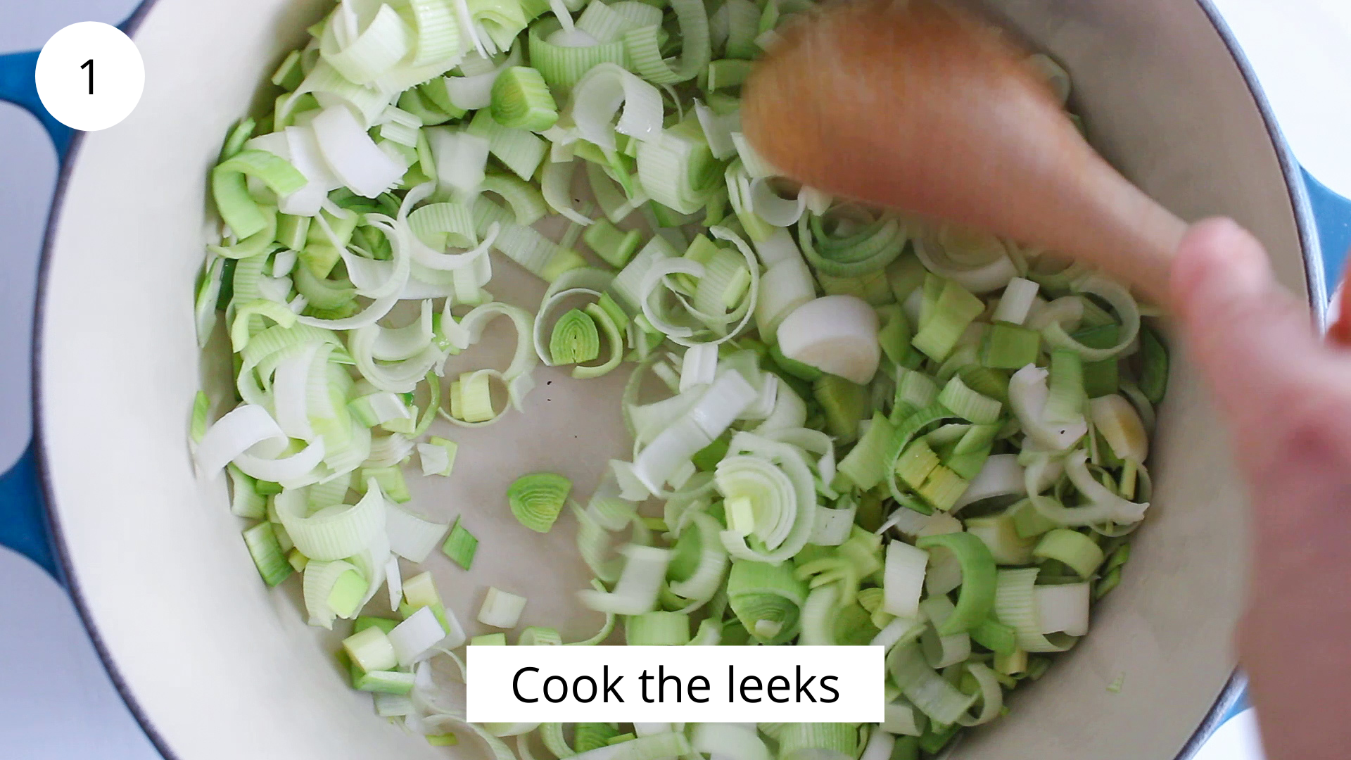 In process picture: Overhead view on a large pot filled with sliced leeks that are cooking with a wooden spoon stirring.