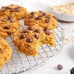 Front angle view on a vegan pumpkin chocolate chip cookie that's placed on a cooling rack and has a bite taken off to show the middle texture. There are rolled oats in the background and chocolate chips in a small bowl as well.