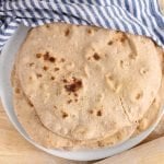 There are a pile of vegan homemade whole wheat tortillas piled on a plate with a damp hand towel over half covering them.