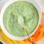 Overhead view on a white bowl that's filled a Vegan Avocado Green Goddess Dressing with a spoon inside and some raw veggies surrounding the bowl.