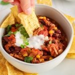 Showing is a tortilla chips that's digging in a bowl of vegan pulled jackfruit chili that's garnished with vegan yogurt and green onion. There are more tortilla chips surrounding the bowl too.