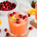 Overhead view showing the top of a glass that has a cranberry, orange and ginger mocktail with some fresh cranberries floating on top of the glass with more fresh fruits in the background.