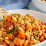 Close up on a vegan American goulash. You can see very well all the lentils and vegetables plus the saucy tomato texture. The dish is garnished with black pepper as well as a side of bread in the background.