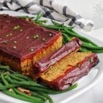 Front view on a vegan lentil mushroom meatloaf with a BBQ tomato glaze that has 2 pieces sliced off with green beans around.