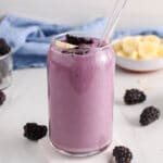 Showing is a glass of strawberry, blackberry and banana smoothie topped with bananas and sliced blackberries and you can see more fruits in the background.