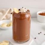 Front angle view on a chocolate banana milkshake. This is a see-through glass so you can see the dark chocolatey drink. Also topped with vegan whipped cream and bananas.